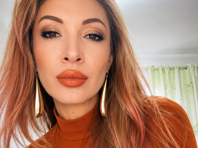 LunaBrice trägt einen orangefarbenen Lippenstift, goldene Ohrringe und einen orangefarbenen Rollkragenpullover. Sie hat braune Augen, volle Augenbrauen und bräunlich-rosa Haare und blickt mit einem neutralen Gesichtsausdruck direkt in die Kamera..