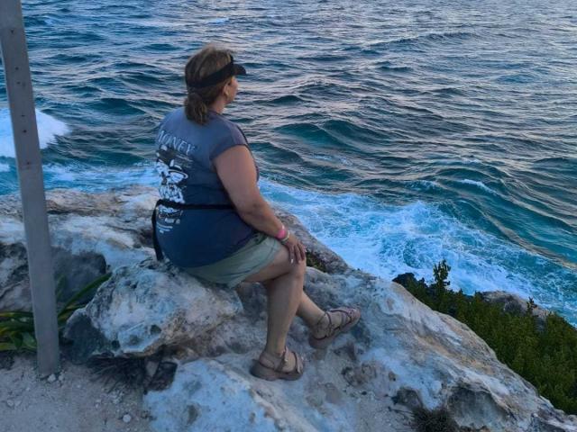 CoupleBomb with brown hair and a visor, wearing a gray shirt and green shorts, sits on rocky shoreline, facing ocean, with blue water and white waves.