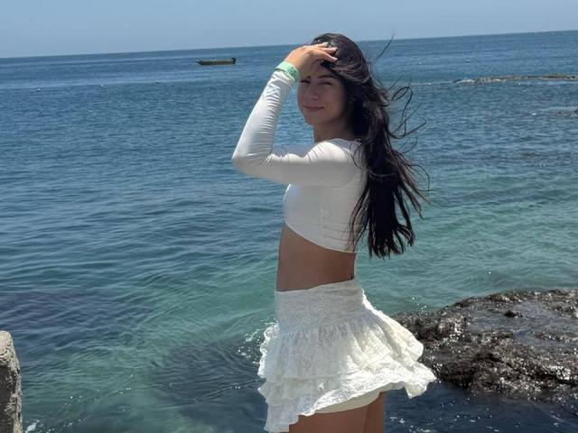 JuliaGarciax, con lunghi capelli scuri, una canotta bianca a maniche lunghe e una gonna bianca in pizzo, in piedi in acqua cristallina blu, con una mano tra i capelli, sorridente, illuminata dal sole, con lo sfondo dell'oceano..
