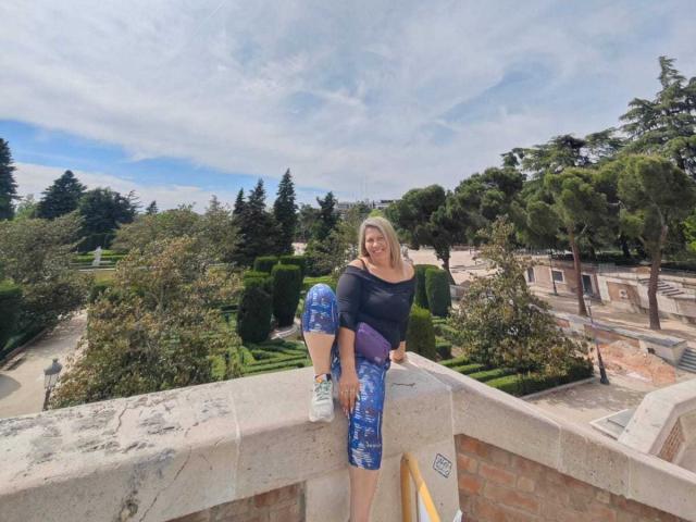 CoupleBomb, a blonde woman with a medium build, is wearing a black top and blue patterned pants, sitting on a stone wall with one leg up, holding a purple bag.