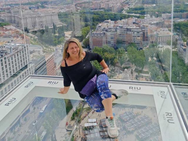 CoupleBomb has blonde hair, black off-shoulder top, blue patterned pants, and white sneakers, sitting on a glass balcony, smiling with arms out, showing her ass and tits.