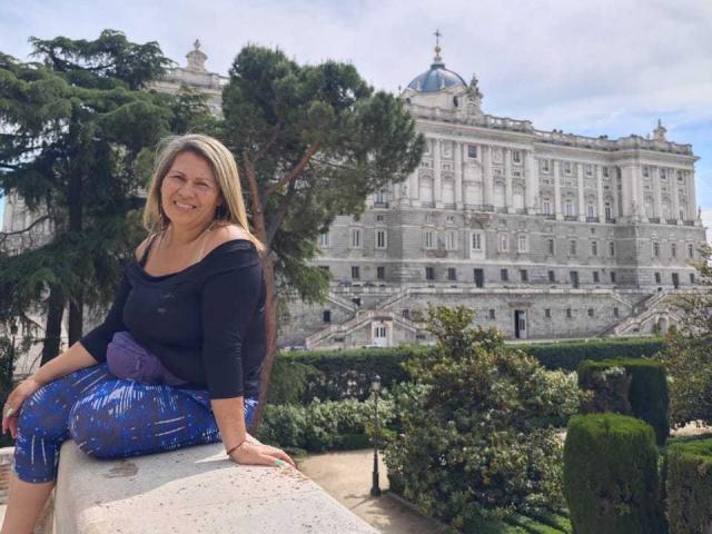CoupleBomb, una donna bionda con un seno di media grandezza, indossa una maglietta nera e pantaloni blu scintillanti, seduta su un muro, sorridente, all'esterno di un grande edificio con una cupola, circondata da alberi e siepi curate..
