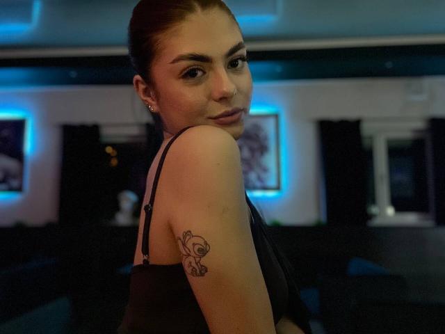 HyonGrillette with brown hair in a black top, looking over her shoulder, showing a tattoo of a cute cartoon girl on her right arm. Bedroom with blue lighting.