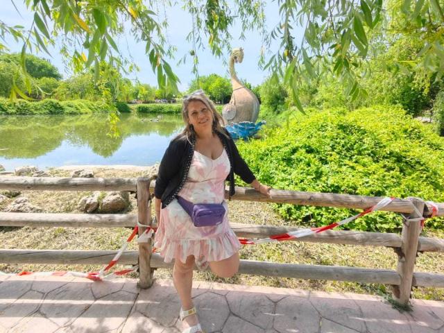 CoupleBomb with blonde hair, pink dress, black cardigan, and purple purse, leaning on wooden fence, smiling, by sunny pond, with statue in background, and hanging green leaves.