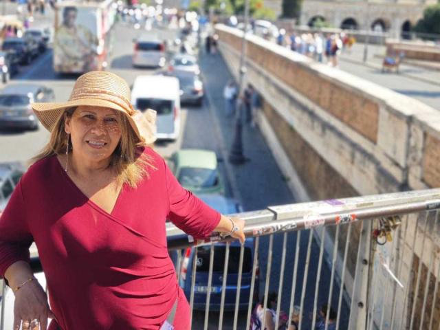 CoupleBomb, una donna di mezza età con un cappello di paglia, una maglia a maniche lunghe rossa e un sedere piccolo, appoggiata a una ringhiera, sorridente, in piedi su un ponte con delle auto sotto..