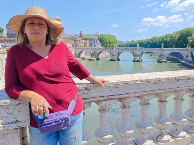 Il sedere paffuto di CoupleBomb, con una maglietta rossa, indossa un cappello di paglia, si appoggia a una ringhiera bianca con una borsa viola, jeans blu e un anello, in piedi vicino a un fiume..