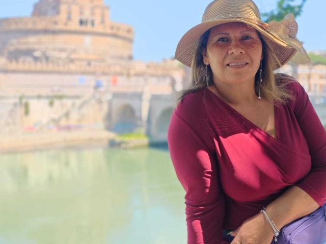 CoupleBomb, middle-aged woman with tan skin, red top, and straw hat, smiling, sitting by green water, showing her tits, wearing purple pants, clear blue sky, sunny day.