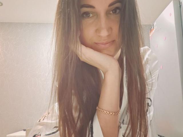 BanyReid, avec de longs cheveux bruns, le menton appuyé sur sa main, portant une chemise blanche avec des motifs noirs et un bracelet en or, souriant doucement à la caméra..