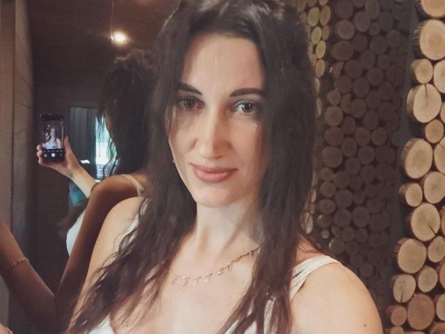 BanyReid, con i capelli castano ondulati, un trucco leggero e un sorriso appena accennato, indossa una canotta bianca e sta scattando un selfie allo specchio, mostrando il riflesso della sua schiena..