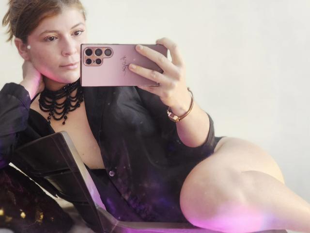 MoonXVelvet macht ein Selfie, trägt einen schwarzen Choker und ein Hemd, zeigt ihre Brüste, sitzt mit angehobenem Bein, hält ein rosa iPhone in der Hand, in einem sanft beleuchteten Raum..