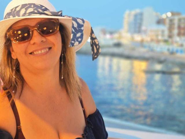 CoupleBomb with sunhat, blue-white pattern, brown sunglasses, smiling, showing boobs in black strap bikini, earrings, blonde hair, by water. Warm sunlight, reflection in water.
