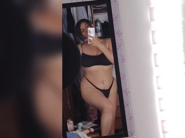 Selfie allo specchio di ZoeyGrey con seno di media grandezza, reggiseno nero e perizoma nero, che tiene in mano il telefono, in piedi in una stanza; il riflesso la mostra sorridente, leggermente formosa, con capelli scuri..