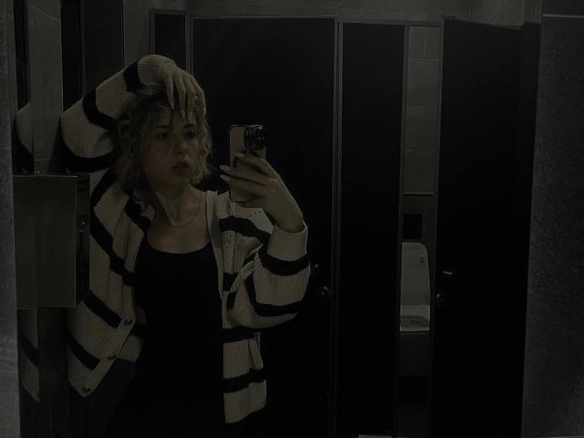 Tora Stambaugh mit welligem, blondem Haar, einem gestreiften Cardigan, einem schwarzen Oberteil, steht in einem dunklen Badezimmer und macht ein Selfie im Spiegel, wobei eine Hand in ihrem Haar ist. Sie sieht ernst aus und hält ein Handy..