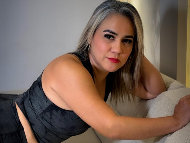CamilaNoer hat mittelgroße Brüste, einen schwarzen Sport-BH und schwarze Hosen, sie lehnt sich an ein weißes Sofa, hat blondes Haar, roten Lippenstift und ein leichtes Lächeln..