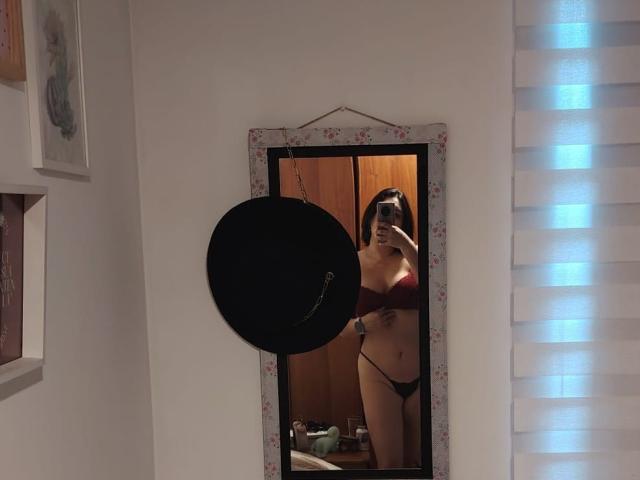 ZoeyGrey stands in front of a mirror, showing off her tits in a red bra and black panties, with a black hat hanging in the way.