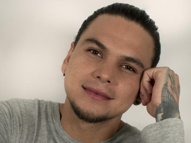 RobSaenz ha una carnagione marrone chiaro, capelli neri raccolti in uno chignon, piccoli tappi per le orecchie neri, una camicia grigia, un tatuaggio sulla mano destra, un sorriso appena accennato, ed ha la testa appoggiata sulla mano..