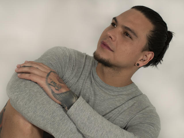 RobSaenz, con i capelli scuri raccolti in uno chignon, indossa una maglietta grigia a maniche lunghe, ha le braccia incrociate mostrando un tatuaggio e dei piercing alle orecchie, guarda leggermente verso l'alto con un'espressione neutra..