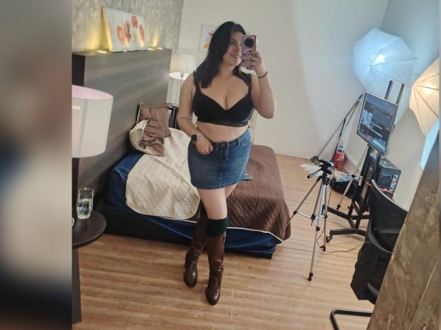 ZoeyGrey with medium tits in a black bra, denim skirt, brown boots, holding phone in mirror selfie, standing in bedroom with bed, lamp, and camera setup.
