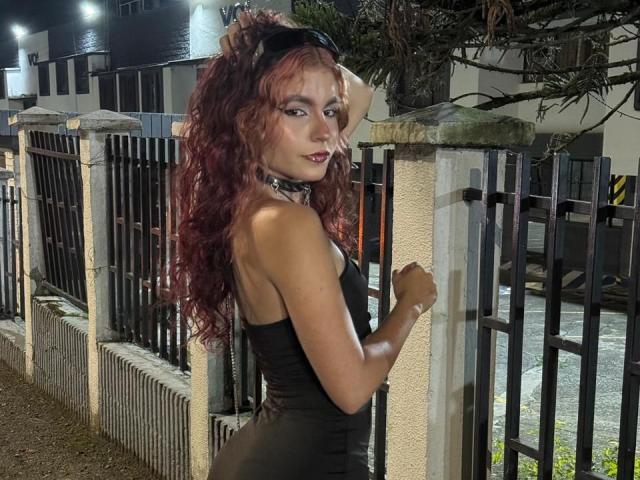 ScarletBlazeX with curly red hair, black strapless leather dress, collar, and sunglasses, standing by a fence at night, looking over her shoulder, showing her ass.