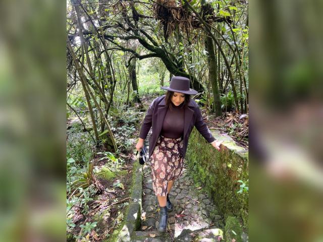 ZoeyGrey with brown hat, dark shirt, floral skirt, black boots, holding camera, walking on mossy stone path in forest, looking down, arms slightly raised.