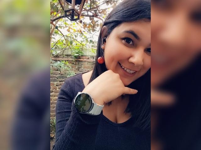 ZoeyGrey with dark hair, red earrings, black sweater, smiling, pointing with her chin up, wearing a green-faced watch, partially blurred face on the right.