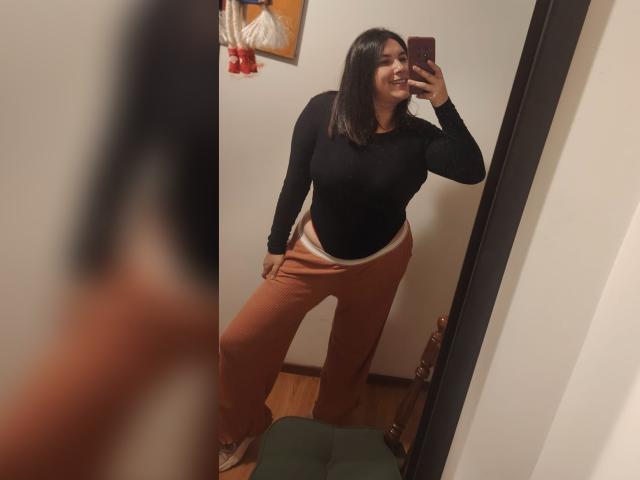 ZoeyGrey, con una maglietta nera, pantaloni marroni e biancheria intima bianca, si scatta un selfie allo specchio, mostrando il sedere, sorridendo, tenendo un telefono, in piedi in una stanza con pavimento di legno..
