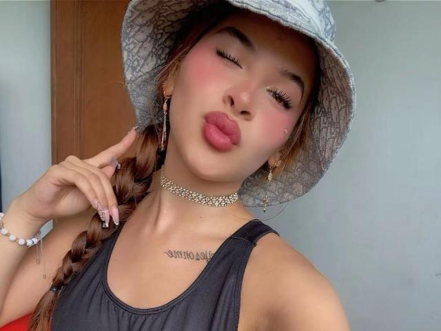 MadisonNoir met volmaakte lippen, bruin gevlochten haar en een grijze bucket hat. Ze draagt een zwarte tanktop, een gouden ketting en heeft een tatoeage onder haar kin..