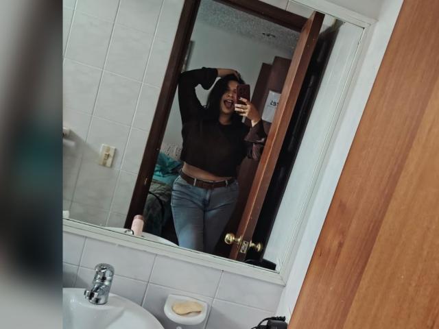 ZoeyGrey è in piedi davanti a uno specchio e sta scattando un selfie. Indossa una maglia nera trasparente che mette in mostra il suo seno, e jeans a vita alta con una cintura marrone..