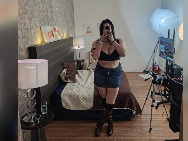 ZoeyGrey è in piedi in una camera da letto, indossa un reggiseno nero, una gonna di jeans, stivali marroni e occhiali, tiene in mano un telefono, mostrando il seno e un leggero girovita, di fronte a uno specchio..