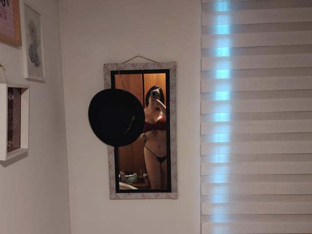 ZoeyGrey standing in front of a mirror, wearing black panties and a red bra, showing her tits and a small tattoo on her right side. A black hat is hanging.