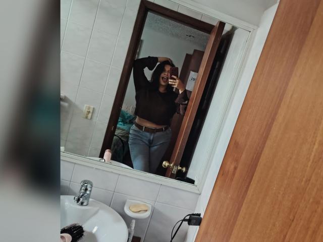ZoeyGrey, con un seno di media grandezza, indossa una maglietta nera trasparente e jeans blu, sta scattando un selfie allo specchio in un bagno, con una mano sulla testa, sorridendo e tenendo il telefono..