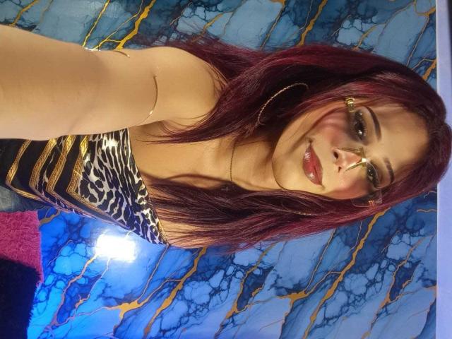 LucianaFlame with red hair, glasses, and hoop earrings, wearing a leopard print top, smiling at the camera, showing off her glossy pink lips and subtle makeup.