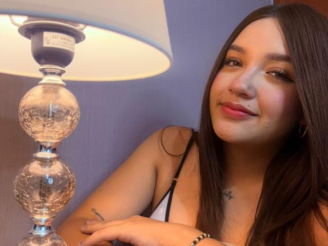 SofiMillerr with long brown hair, smiling, wearing a black spaghetti strap top, next to a clear, textured lamp with a white lampshade. Her tits are small and she's cute.
