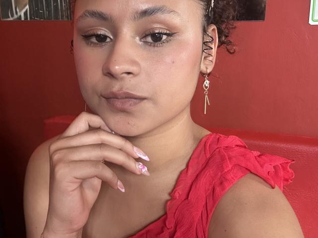 Sofia, met krullend bruin haar, lichtbruine huid, een rode jurk, roze nagellak en lange oorbellen, die haar kin aanraakt, kijkt naar de camera, met een rode muur op de achtergrond..