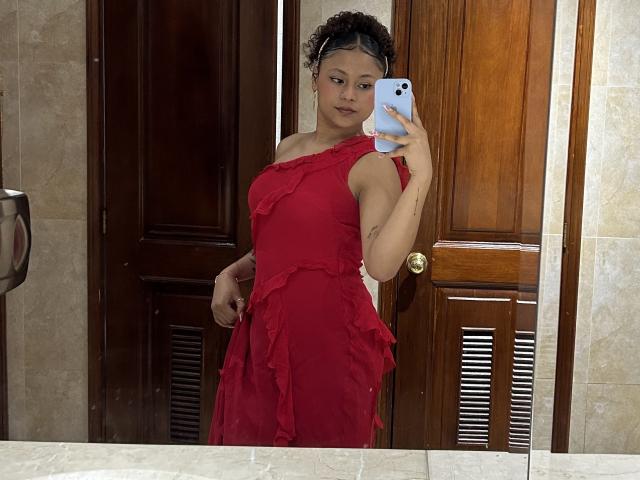 Sofia, com cabelos castanhos cacheados, vestindo um vestido vermelho com babados, segurando um telefone, tirando uma selfie no espelho, mostrando seus seios e uma pequena tatuagem no braço direito..