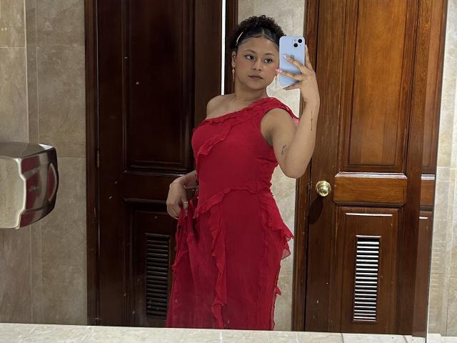 Sofia, passionnée, vêtue d'une robe rouge à volants, tenant un téléphone, debout devant une porte en bois, avec une expression sérieuse, des seins de taille moyenne, des cheveux foncés et une peau claire..