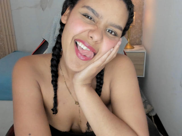AriaVega, con cabello negro trenzado, sonriendo con la lengua afuera, mano en la mejilla, mostrando senos pequeños, piel de color marrón claro, usando un collar, en un dormitorio con una lámpara..