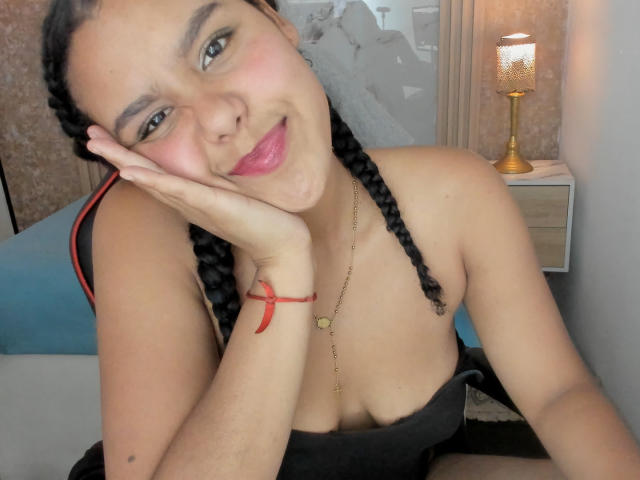 AriaVega, con cabello negro trenzado, labios rosados y un collar de oro, apoyada en su mano, sonriendo a la cámara, mostrando sus senos, en un dormitorio con una lámpara..