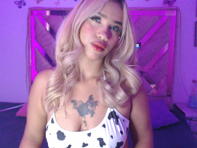 LuluDeVine, con rizos rubios, lápiz labial rojo y una blusa de vaca en blanco y negro, mostrando sus pechos y un tatuaje sobre su pecho, en un dormitorio iluminado con luz púrpura..