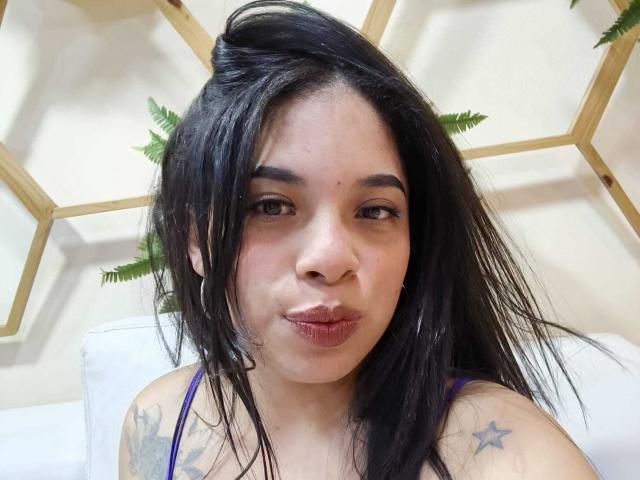 SammyRossek with puffy lips, black wet hair, purple top, star tattoos, and hoop earrings, making a pouty face. Her brown eyes and subtle makeup are visible.