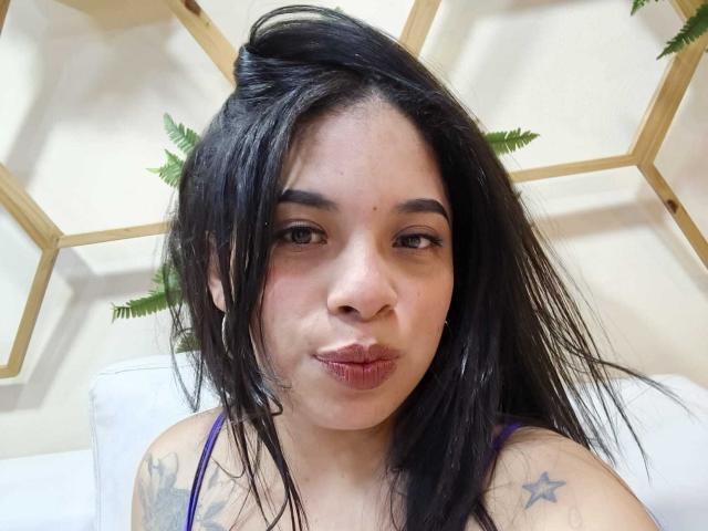 SammyRossek with black hair, pouty lips, brown eyes, wearing a purple top, showing tattoos on her shoulder, and hoop earrings, looking at the camera.