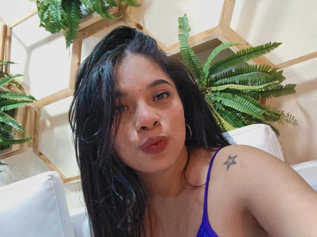 SammyRossek with black hair, purple bra, hoop earrings, star tattoo on shoulder, pouty lips, green ferns in background, wooden hexagon wall, white couch, natural light.