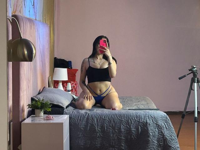 TanyaEmas, kneeling on a bed with a grey quilt, wearing a black crop top and blue thong, showing her tits and pussy, holding a red phone.