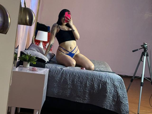 TanyaEmas with tattoos, black crop top, blue strappy panties, kneeling on gray bed, holding pink phone, mirror selfie, wooden floor, tripod on right, bedside lamp on left.