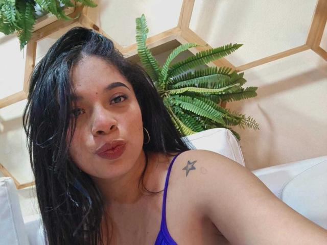 SammyRossek, con capelli scuri e ondulati, reggiseno viola, orecchini a cerchio, tatuaggio a forma di stella sulla spalla destra, labbra carnose e pelle marrone, seduta su un divano bianco con una felce verde sullo sfondo..