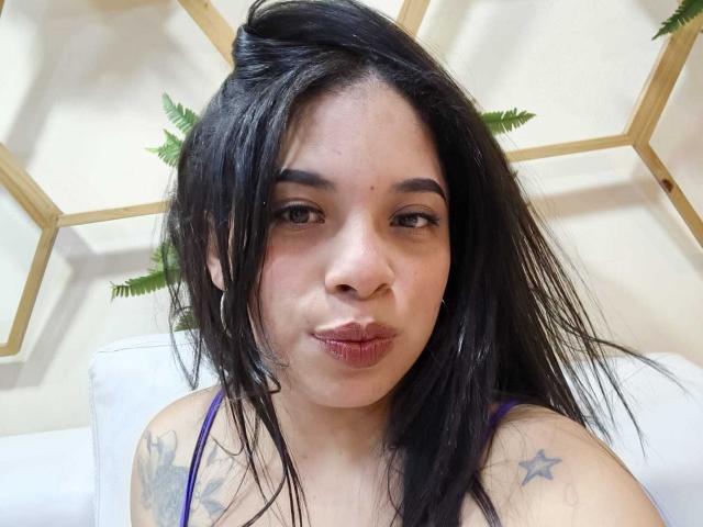 SammyRossek, con i capelli neri bagnati, labbra rosse e gonfie, occhi marroni, piccoli orecchini a cerchio, top viola, tatuaggi sulla spalla, che fa una smorfia, guardando la fotocamera..
