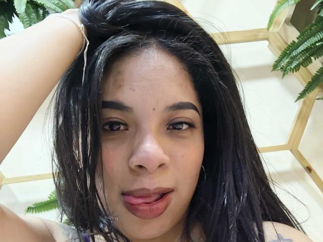 SammyRossek tem cabelo escuro e molhado, uma mão levantada, olhos castanhos e uma pequena pinta no nariz. Ela está com a língua rosa para fora. As nádegas dela não estão visíveis..