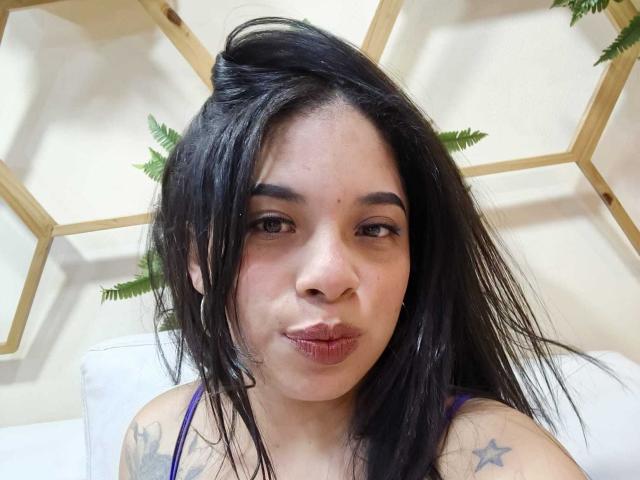 SammyRossek, con i capelli scuri e lucidi, labbra piene, piccoli orecchini a cerchio e tatuaggi a forma di stella sulle spalle, guarda direttamente l'obiettivo..