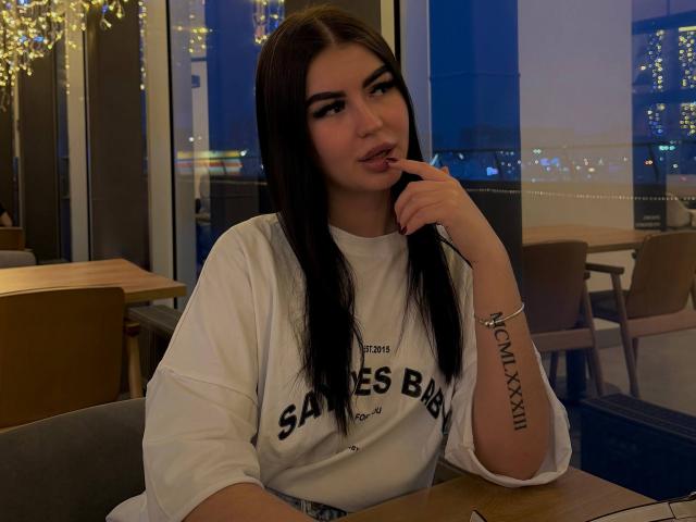 LeonaMeilee mit langen, schwarzen Haaren, die ein weißes "Sances Bay"-Shirt trägt, beißt sich in den Finger, hat ein Tattoo am rechten Unterarm und sitzt in einem schwach beleuchteten Restaurant und schaut in die Kamera..