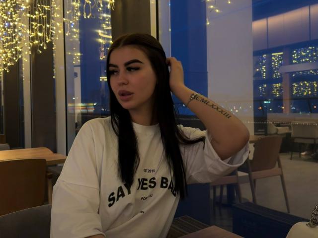LeonaMeilee hat dunkles Haar, ein Tattoo auf ihrem rechten Unterarm und trägt ein weißes T-Shirt mit der Aufschrift "SAV JES BANQ". Sie hat ihre Hand im Haar und schaut in die Kamera..